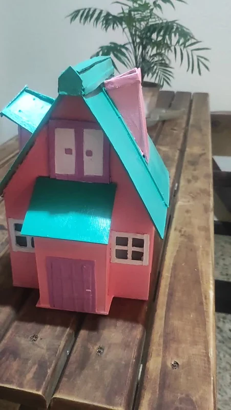 Casinha de boneca rosa, com telhado verde-água e janelas em relevo, sobre uma bancada de madeira